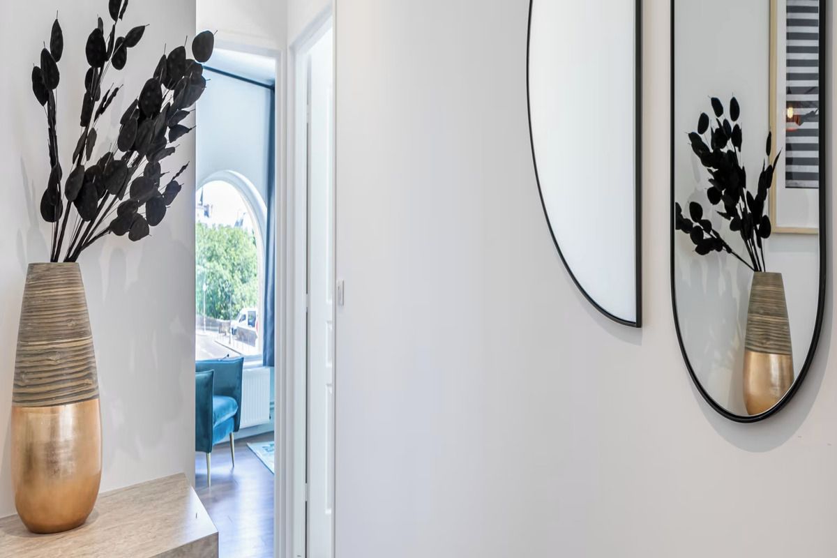 A modern interior with a vase of black leaves on a wooden surface, a mirror on the wall, and a glimpse of a blue chair and a door leading outside.