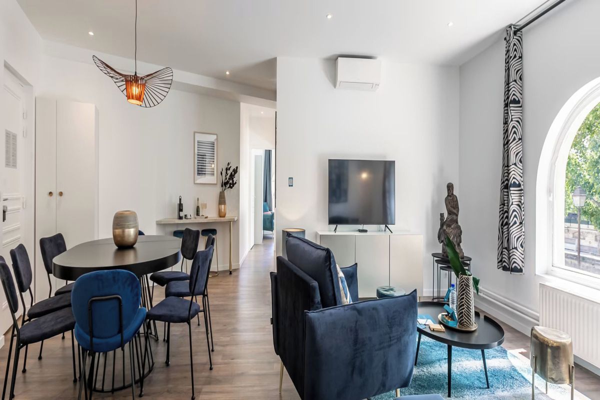 A modern living room with a dining area, a flat-screen TV, and a large window with curtains.