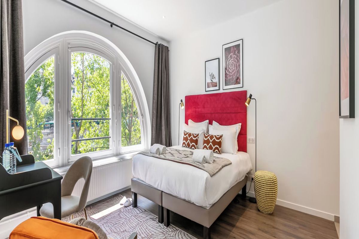 Een moderne slaapkamer met een groot raam, een bed met een rode hoofdeinde, een bureau en een stoel.