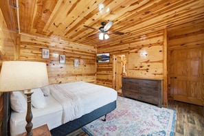 A wooden cabin bedroom with a bed, a dresser, a ceiling fan, and a television on the wall. - Make Lasting Memories: Pool, Hot Tub, Games & MORE (Pigeon Forge)