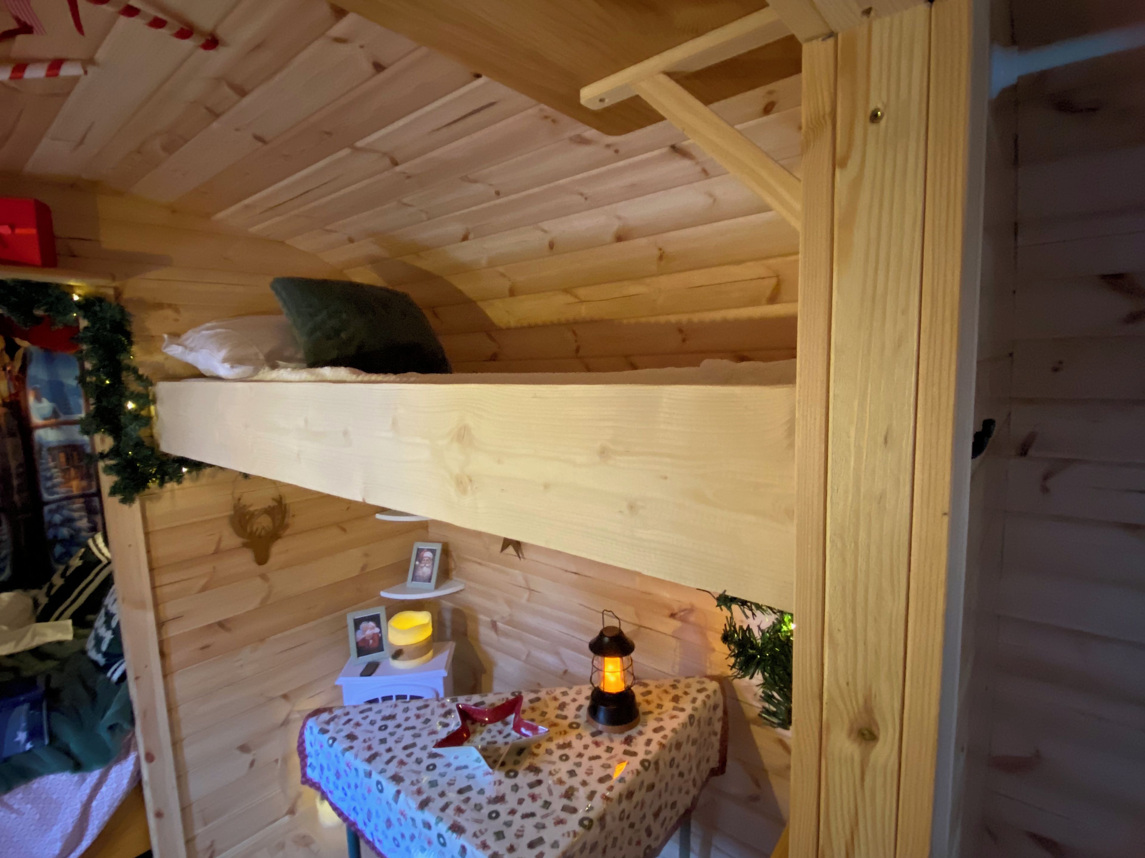 A wooden bunk bed with a ladder, a small table with a patterned tablecloth, a lantern, and a Christmas tree decoration.
