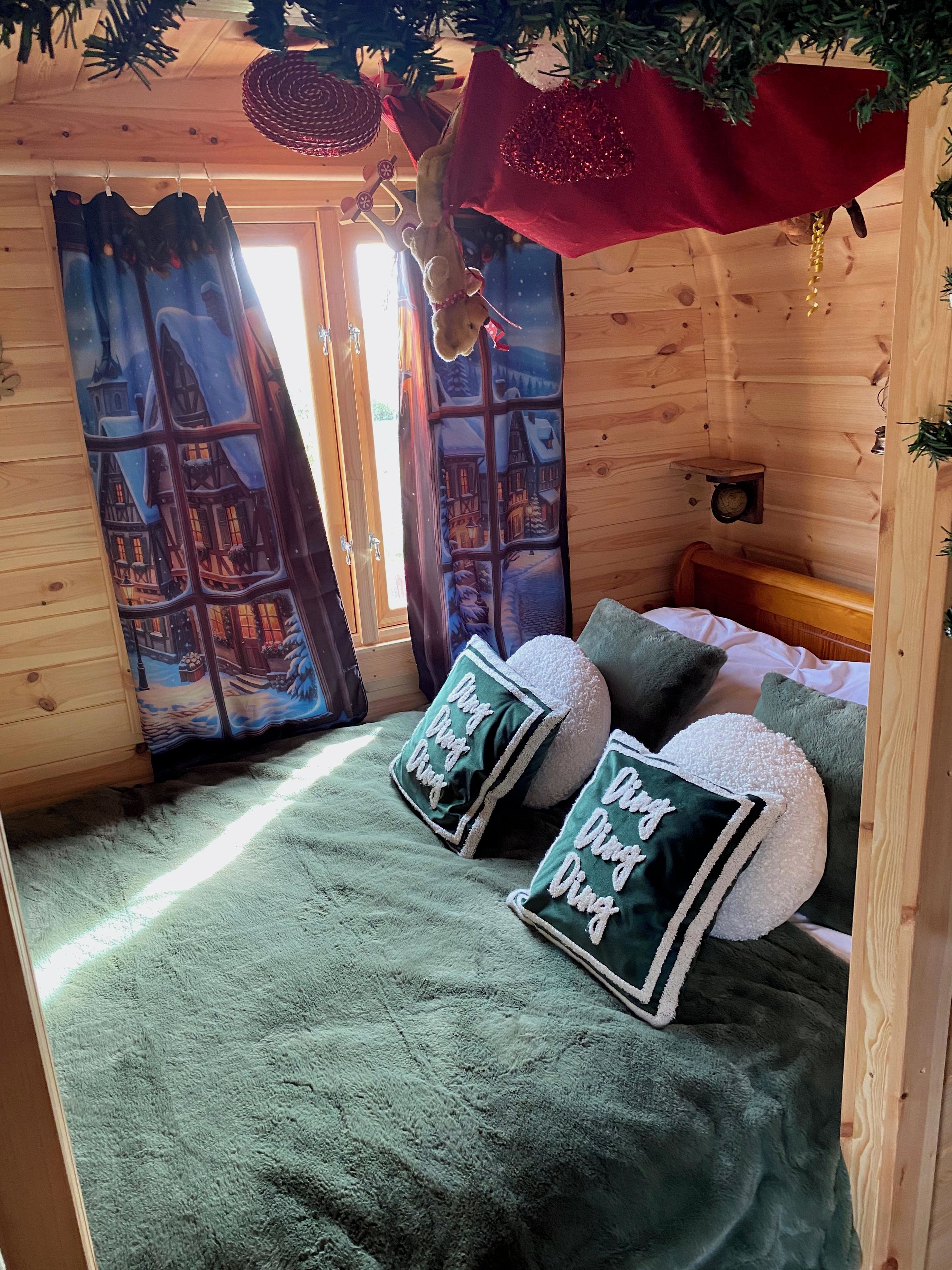A cozy room with a bed, green pillows, a window with a view of a snowy village, and a Christmas tree.