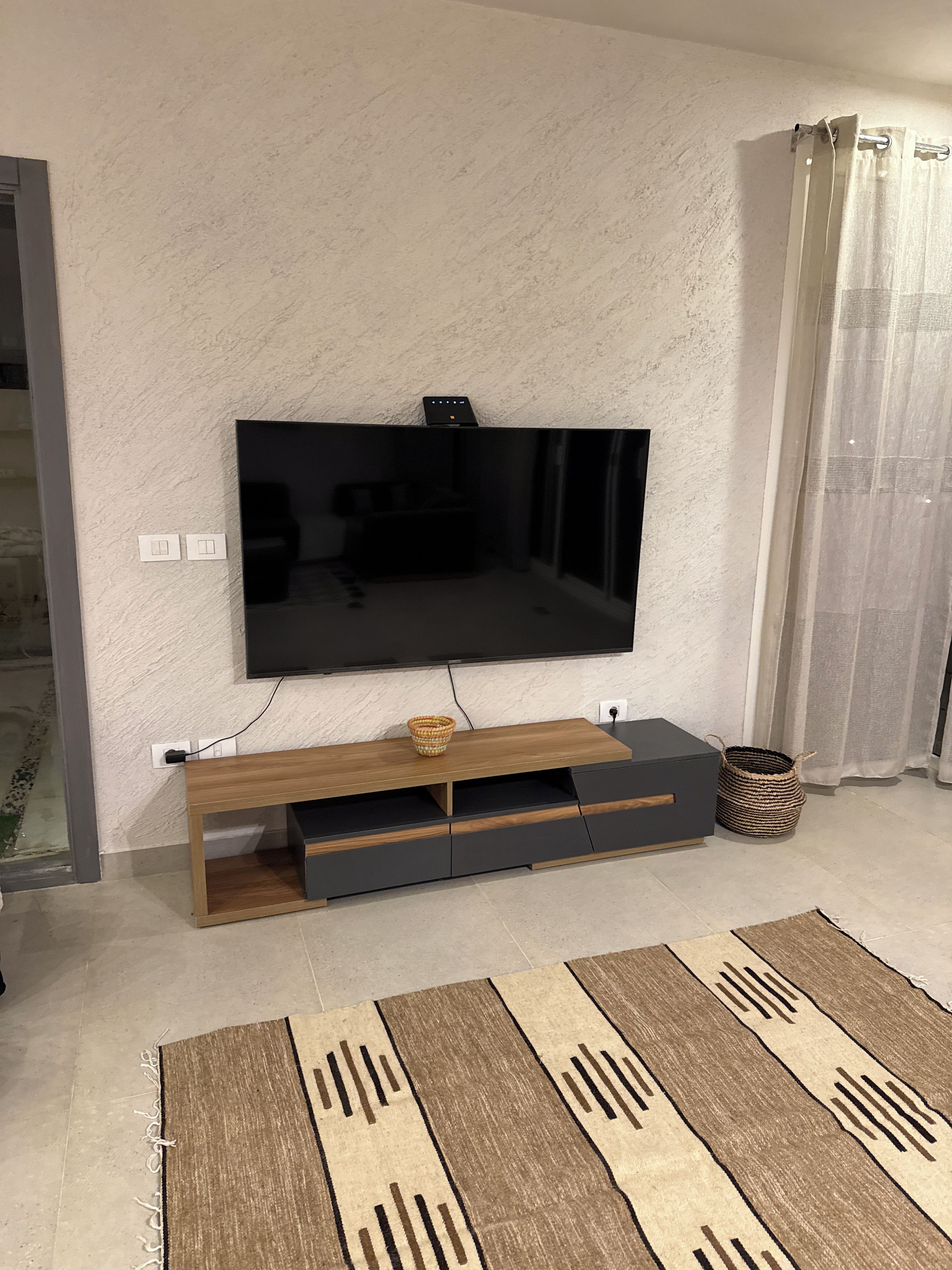 A flat-screen TV mounted on a textured wall, a wooden TV stand with drawers, a woven basket, and a patterned rug on a tiled floor.