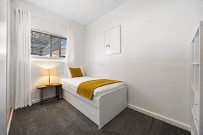 A small, single-bed room with a window, a bedside table with a lamp, and a framed picture on the wall. - Glee Tides By the Beach Indoor outdoor Lifestyle (Glenelg East)