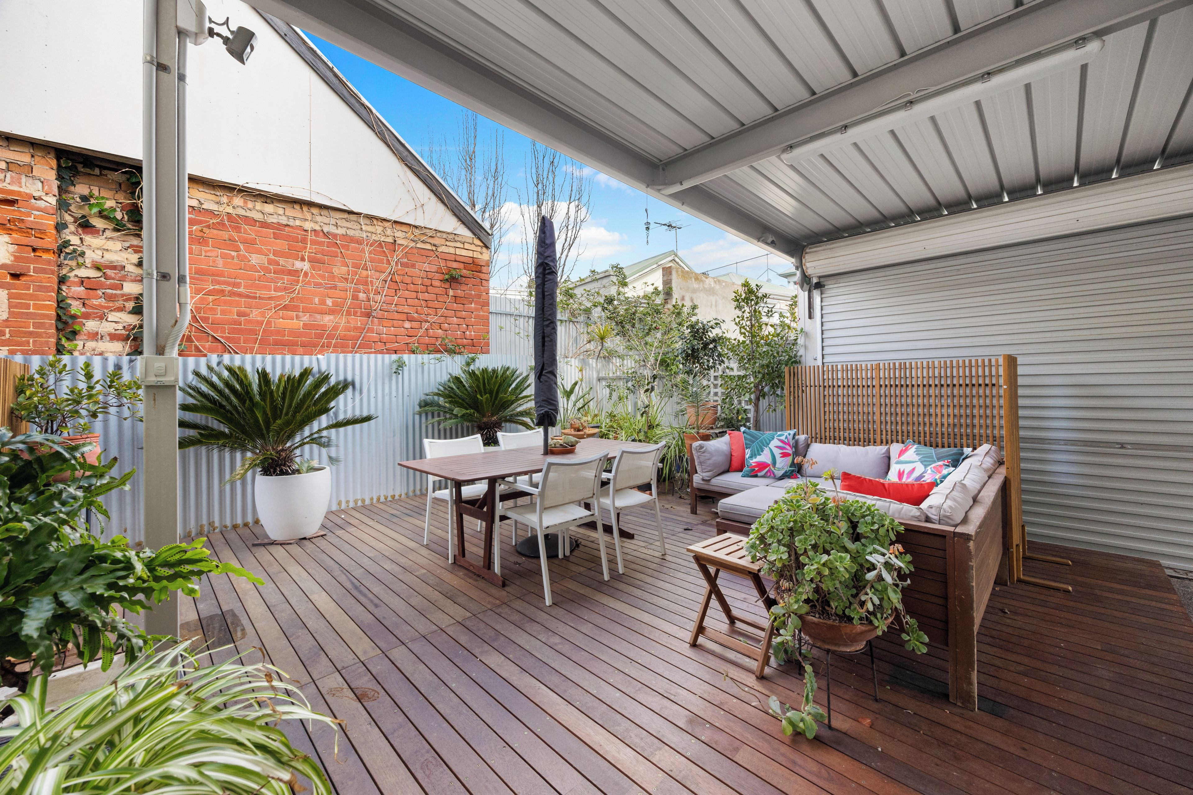 A covered outdoor patio with a wooden deck, a dining table with chairs, a sofa with cushions, and various potted plants.