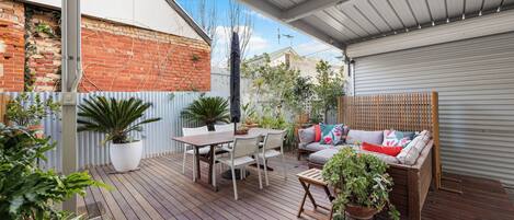 A covered outdoor patio with a wooden deck, a dining table with chairs, a sofa with cushions, and various potted plants.