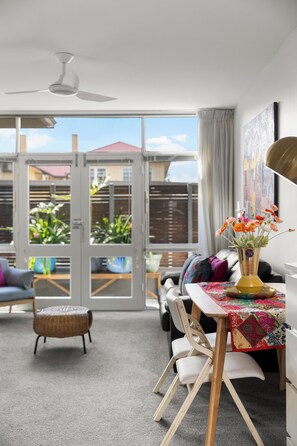 A modern living room with a dining area, a ceiling fan, a large window with a view of the outside, and a table with a colorful tablecloth. - Glee Tides By the Beach Indoor outdoor Lifestyle (Glenelg East)