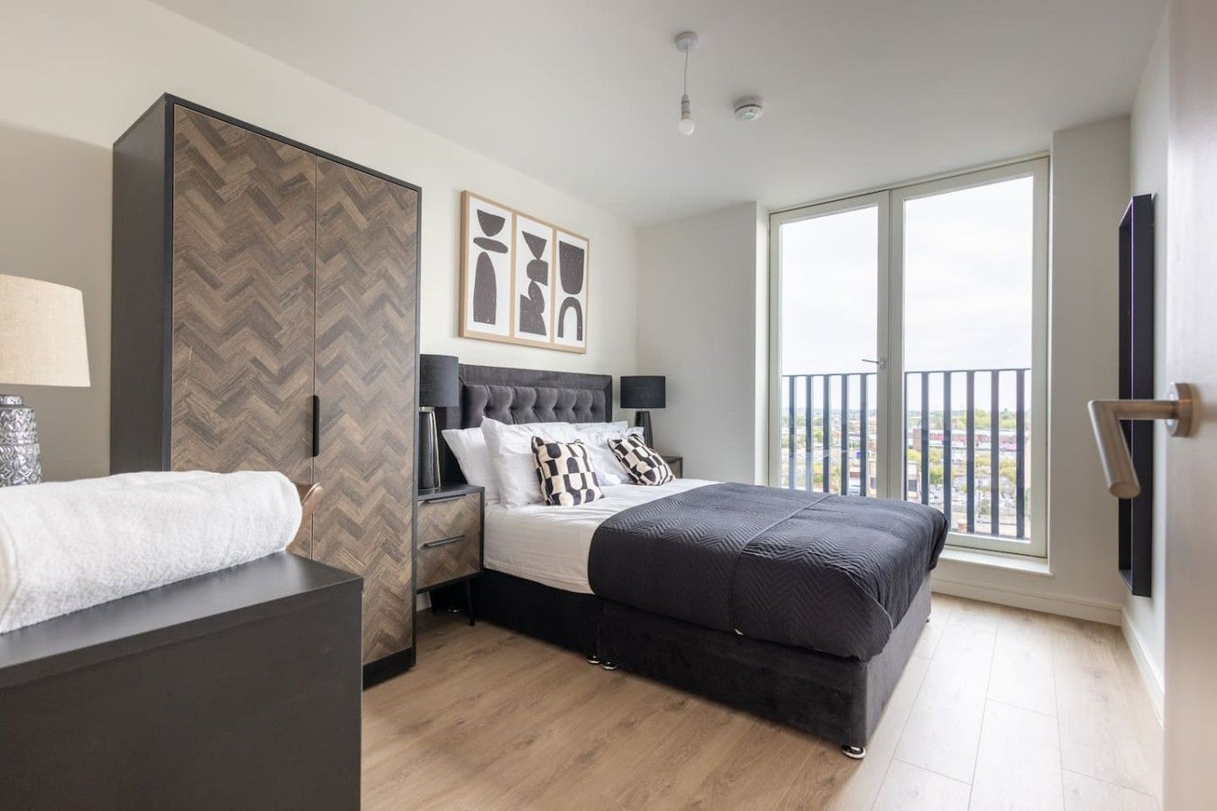 A modern bedroom with a large bed, a wardrobe, a nightstand, and a window with a balcony view.