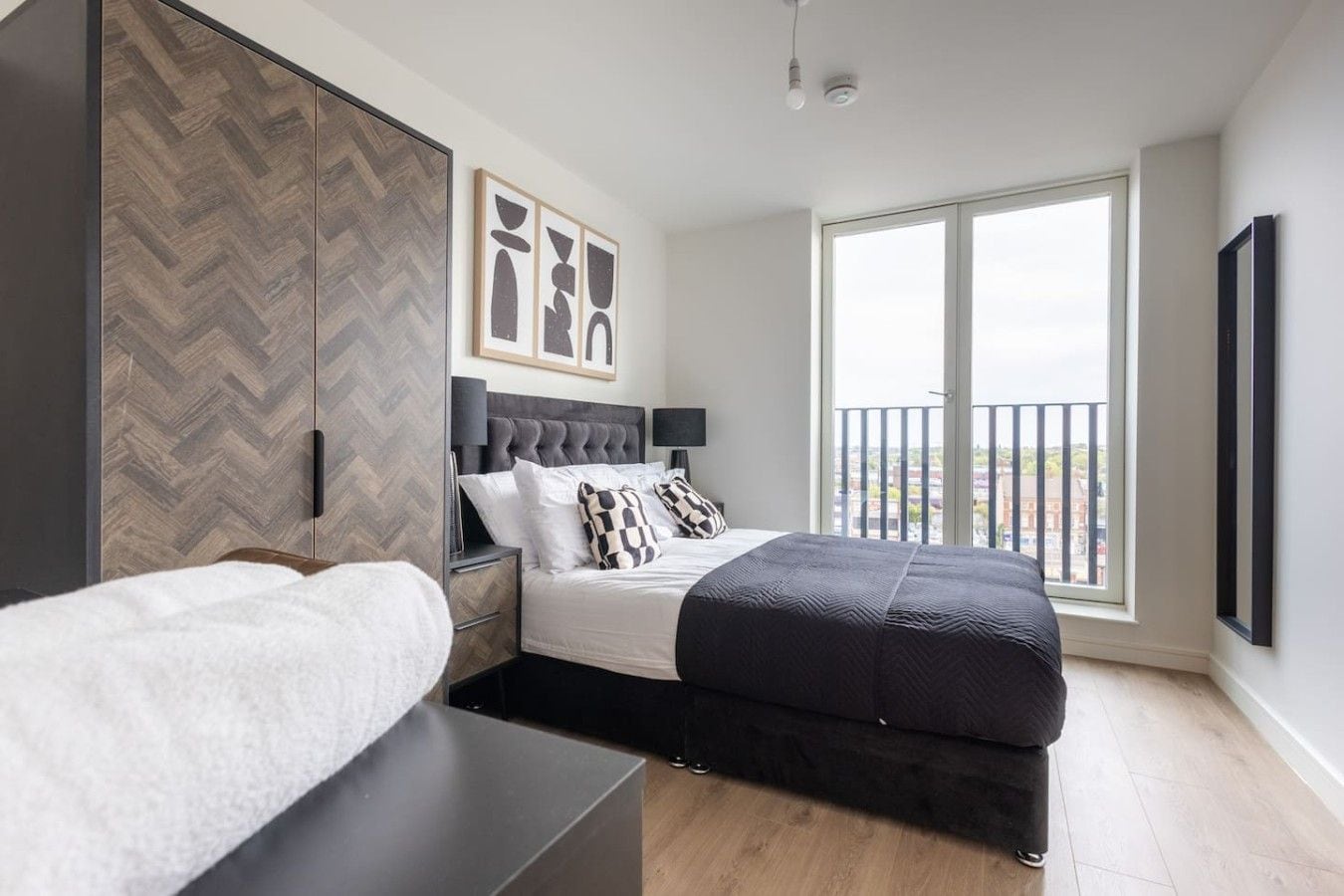 A modern bedroom with a bed, bedside tables, a wardrobe, and a large window offering a city view.
