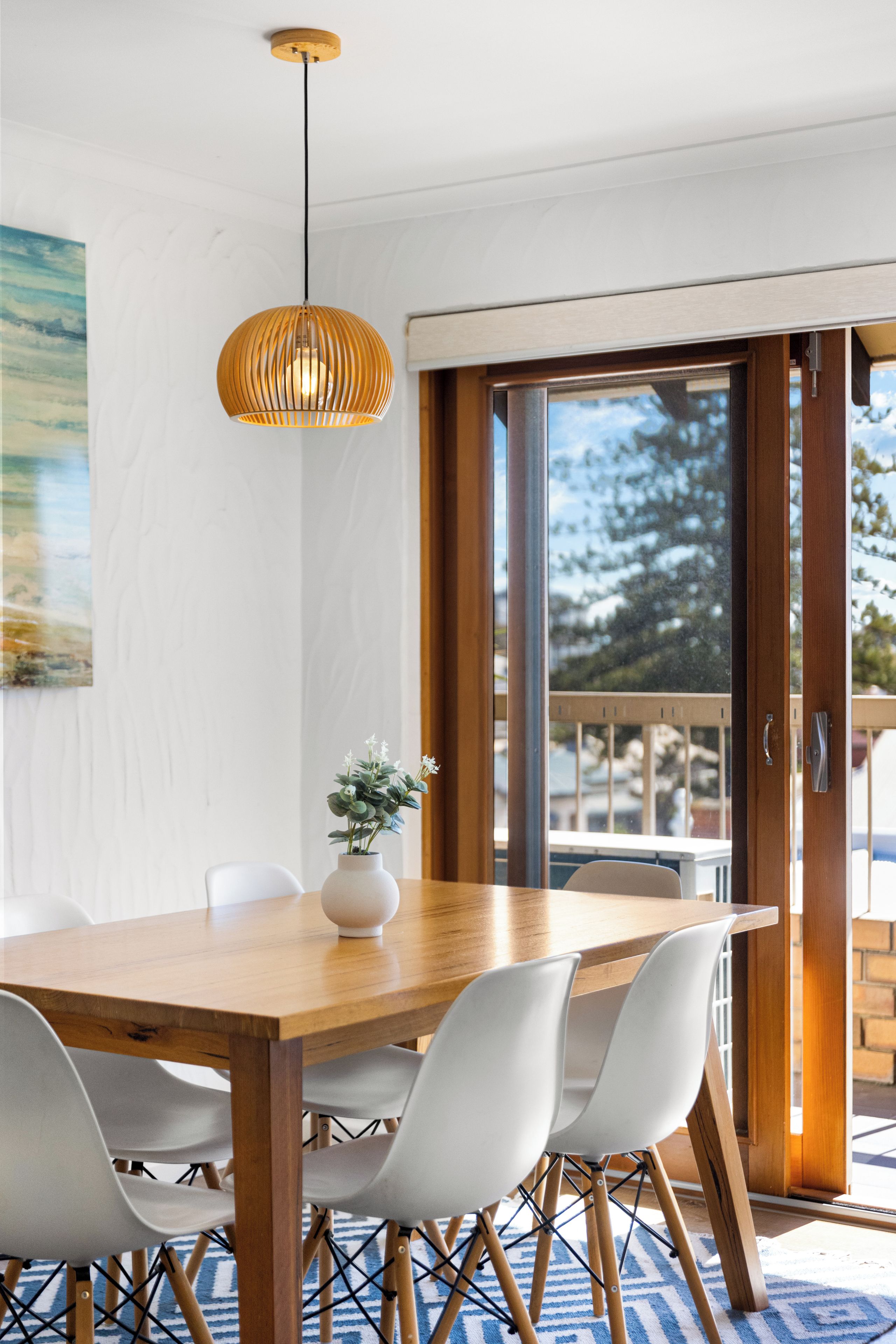 A dining area with a wooden table, white chairs, a hanging pendant light, and a vase with flowers.