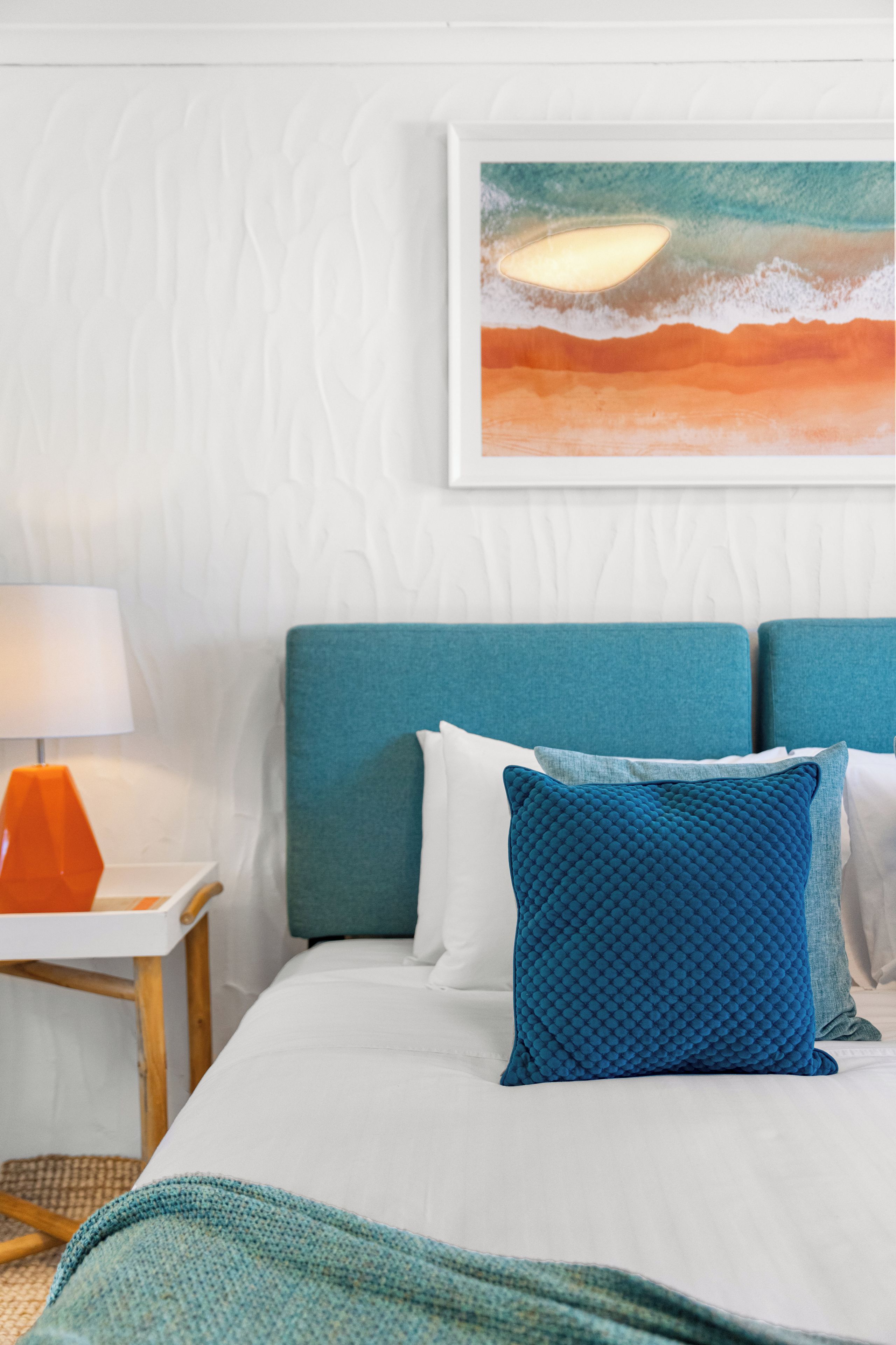 Habitación de hotel con cama, almohadas, mesita de noche, lámpara y un cuadro colgado en la pared.