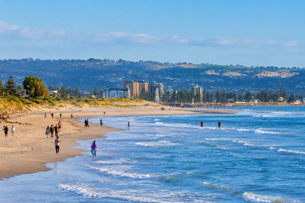 A beach with people walking along the shore, buildings in the background, and a hilly landscape.