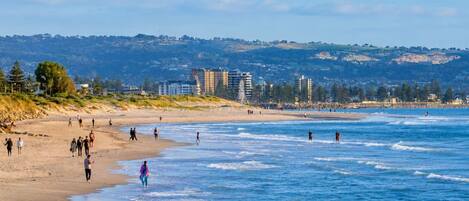 A beach with people walking along the shore, buildings in the background, and a hilly landscape.