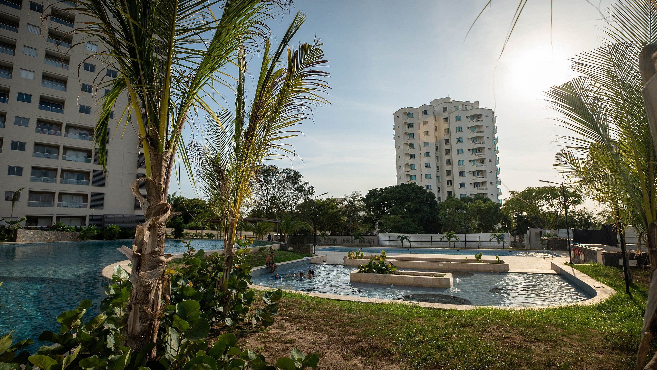 Zona de piscina con múltiples fuentes, rodeada de palmeras y un edificio residencial alto al fondo.