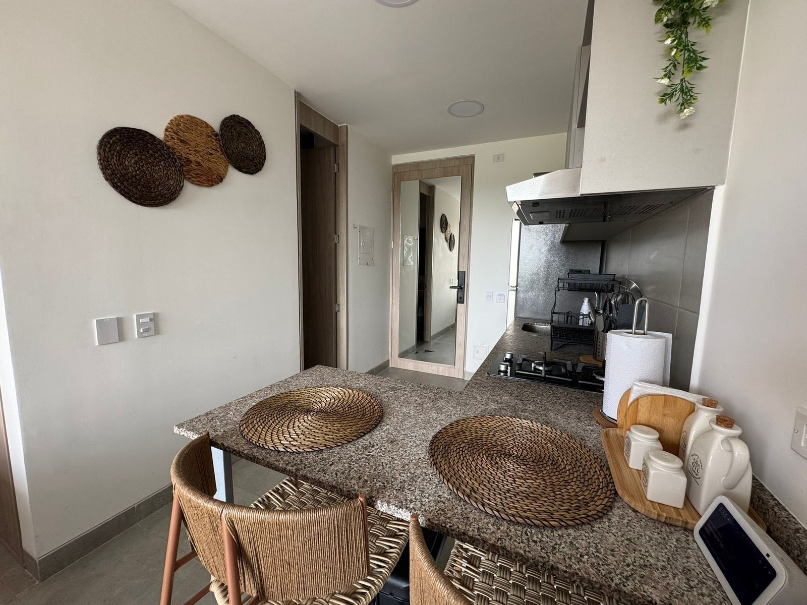 Una cocina moderna con encimeras de granito, horno empotrado y campana extractora de acero inoxidable. Hay dos individuales tejidos sobre la mesa, y un pequeño dispositivo digital en la encimera.