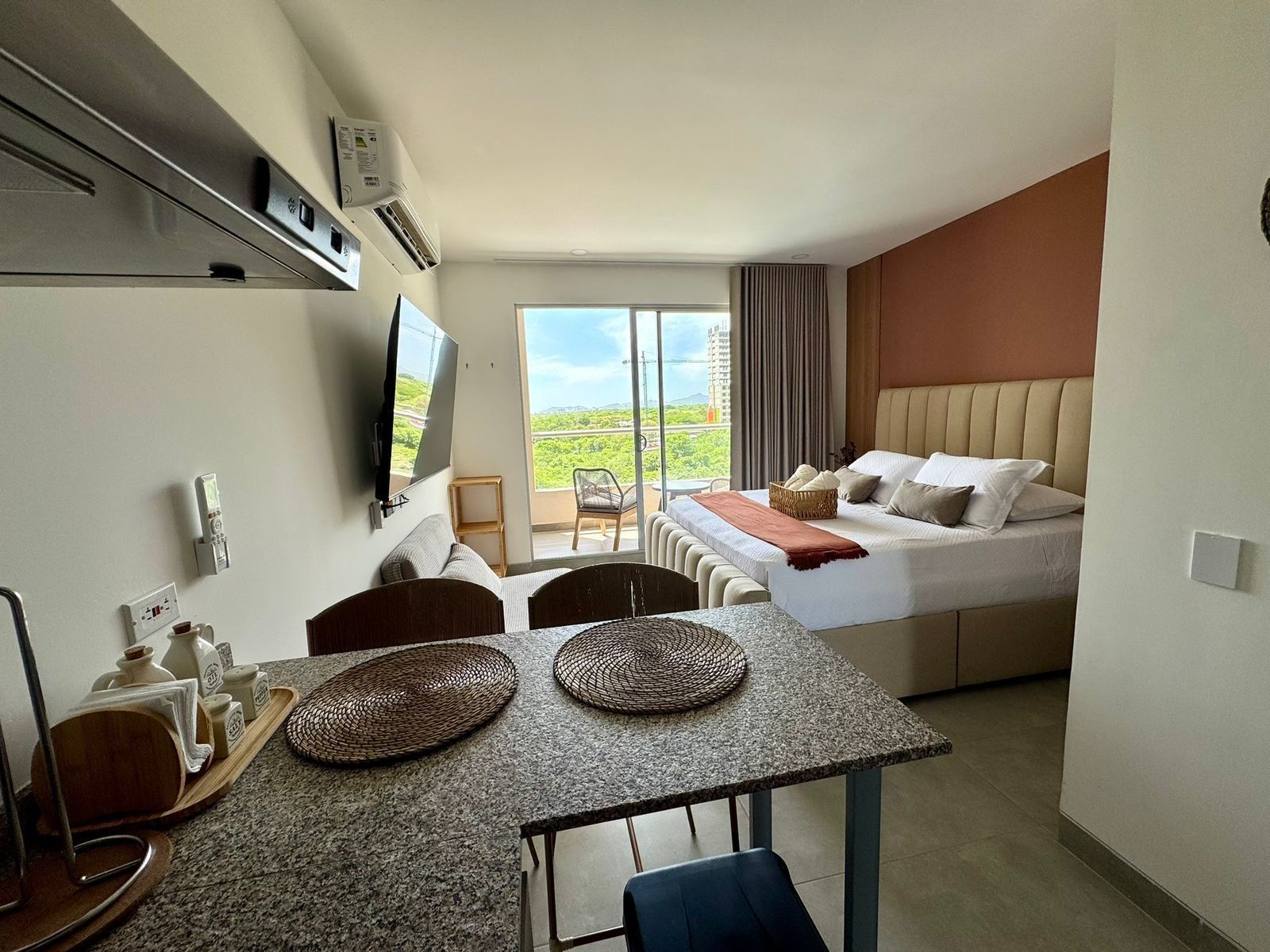 Quarto de hotel com cama, mesa de jantar com cadeiras, televisão na parede e uma grande janela com vista para o exterior.