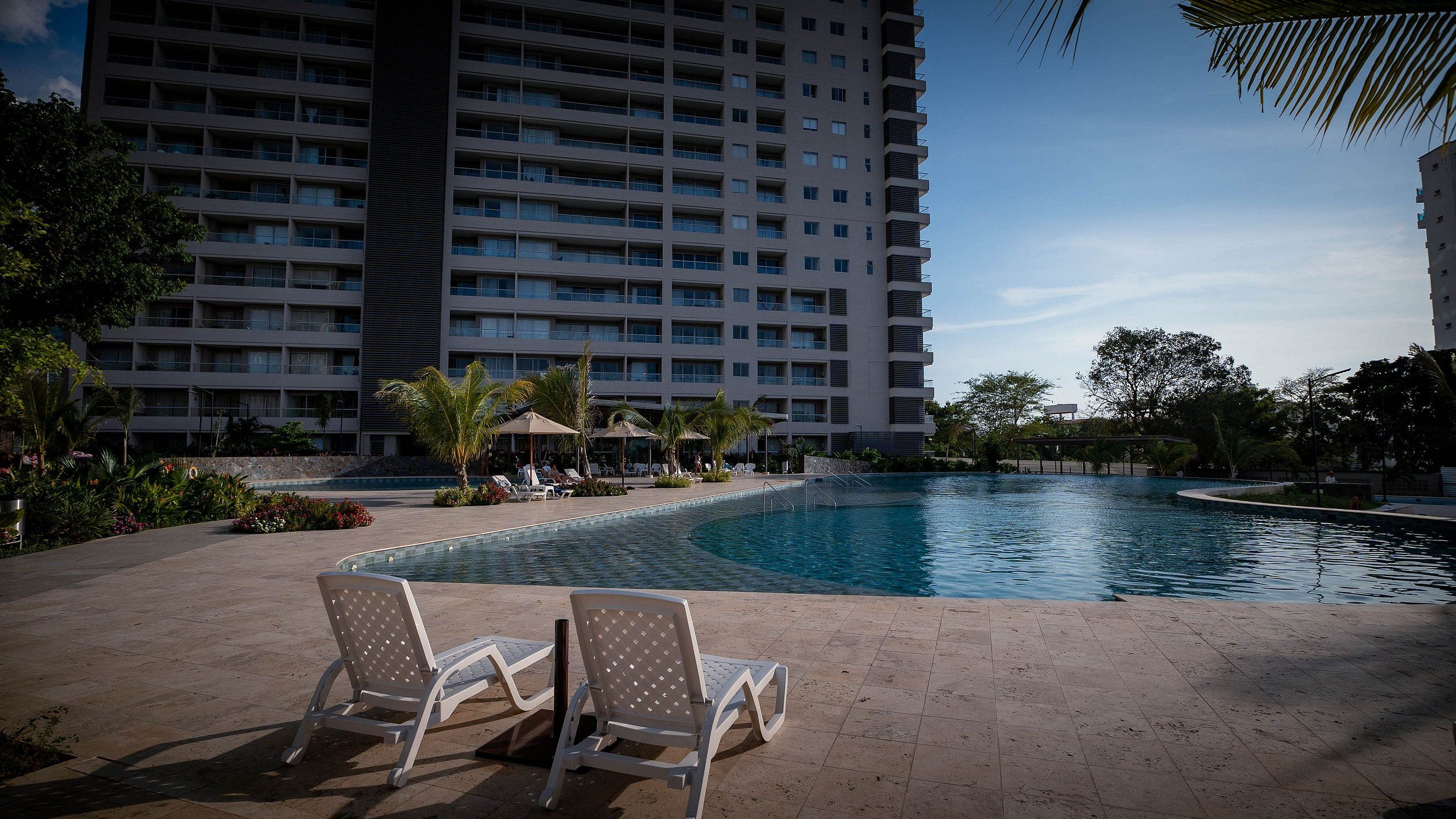 Une piscine entourée de chaises longues, elle-même bordée d’un immeuble doté de nombreux balcons.