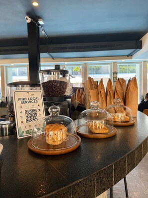 A café counter with a sign for local services, a coffee bean dispenser, and pastries under glass cloches. - Upscale Playacar Studio w Beach Club & 4 Pools (Playa del Carmen)