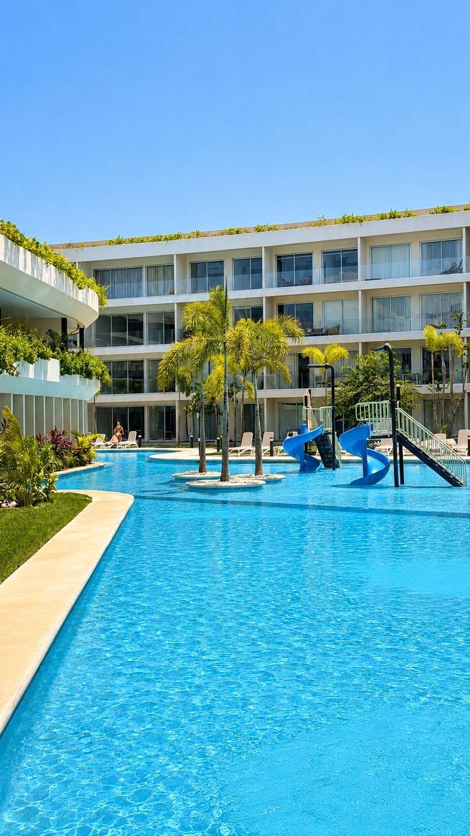 En swimmingpool med rutschkanor, omgiven av en byggnad med balkonger och grönska.