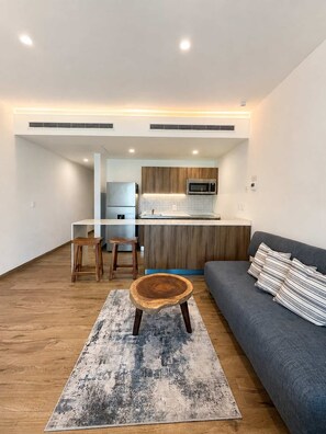 A modern living room with a grey sofa, a wooden coffee table, and a kitchen area with a microwave and refrigerator. - Upscale Playacar Studio w Beach Club & 4 Pools (Playa del Carmen)