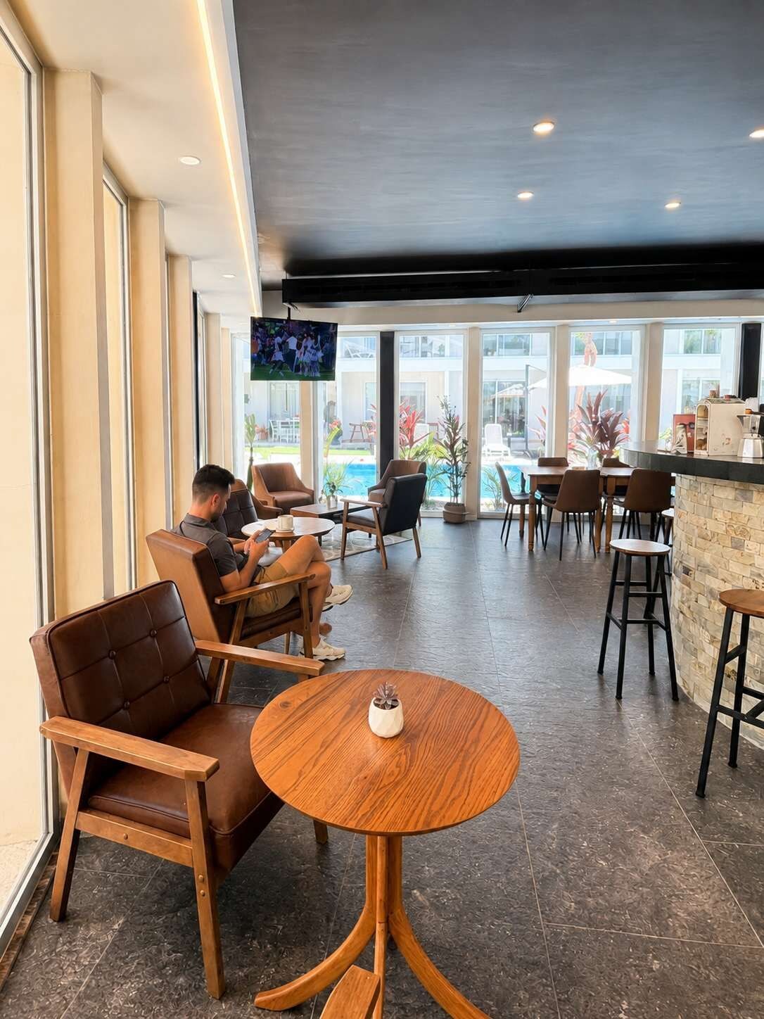 A modern indoor seating area with leather chairs and a round wooden table, overlooking a pool and outdoor seating.