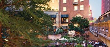 A riverside courtyard with outdoor seating, a brick building, and a bridge.