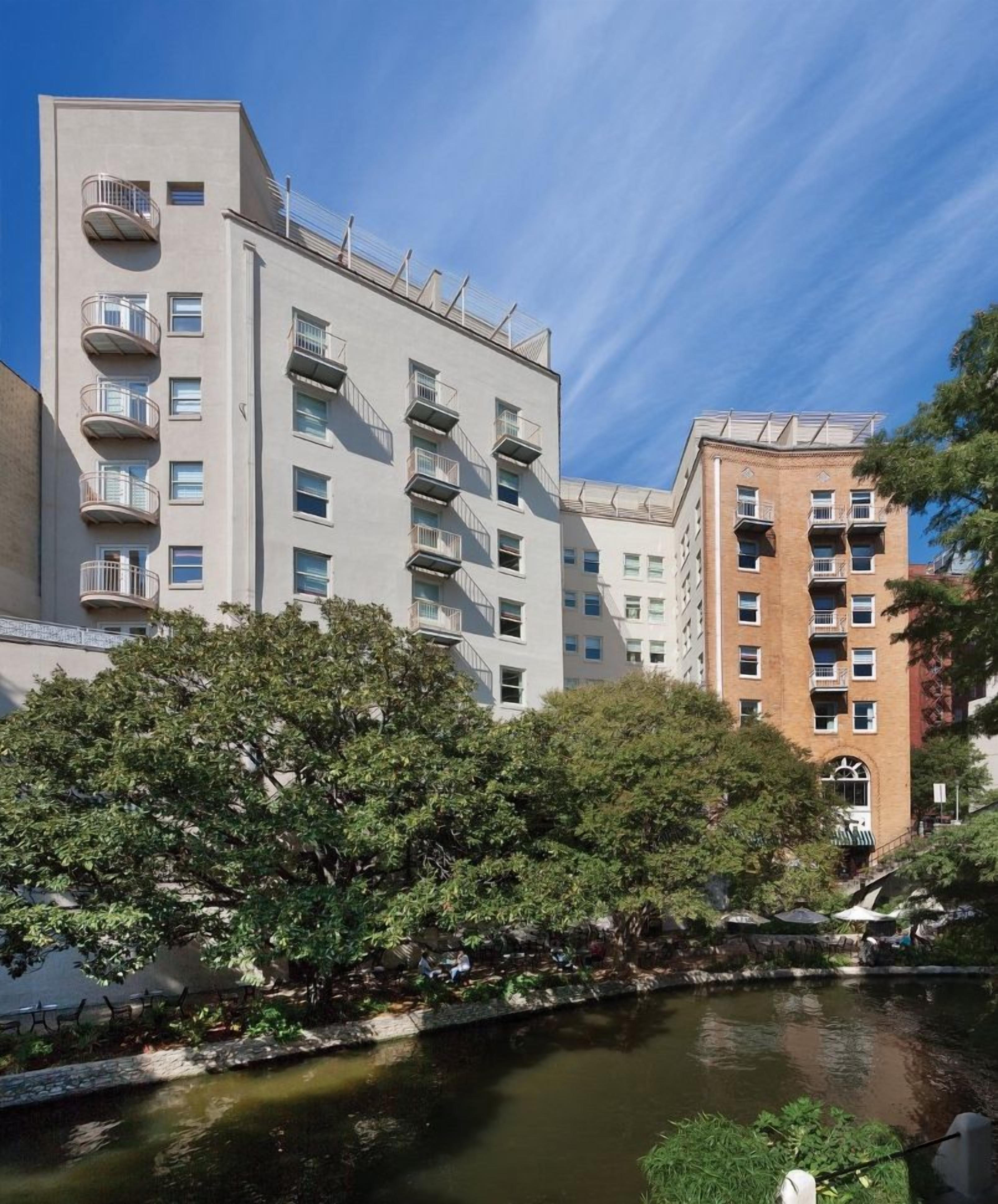 A multi-story building with balconies, a river in the foreground, and trees.