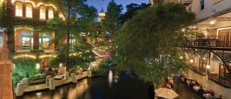 A riverside walkway with outdoor seating, a bridge, and buildings with illuminated windows.