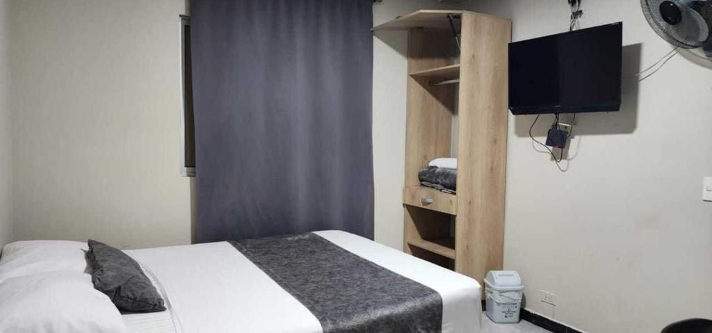Un lit simple avec une couverture et des oreillers gris, une armoire en bois, un téléviseur fixé au mur et une petite poubelle.