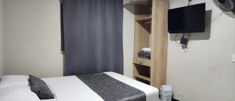 A single bed with a grey blanket and pillows, a wooden wardrobe, a television mounted on the wall, and a small trash bin.