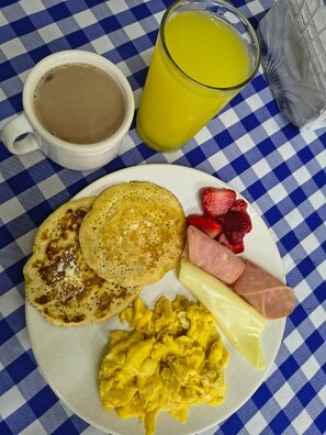 Un plato de desayuno con tortitas, huevos revueltos, salchichas, fresas y un vaso de zumo de naranja.