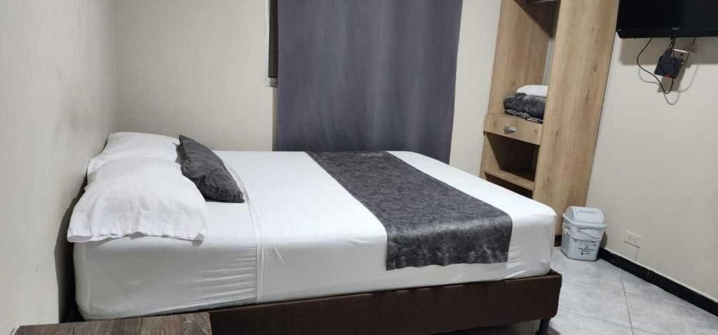 Uma cama de solteiro com colchão branco, cobertor cinza e travesseiro. Possui cabeceira de madeira, prateleira pequena com um cobertor dobrado e uma lixeira à direita.