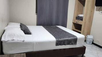 Uma cama de solteiro com colchão branco, cobertor cinza e travesseiro. Possui cabeceira de madeira, prateleira pequena com um cobertor dobrado e uma lixeira à direita.