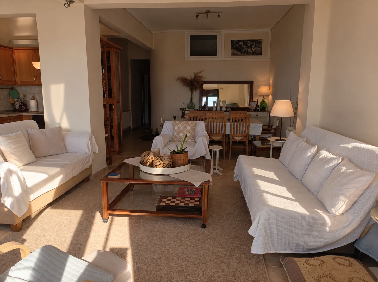A living room with two white sofas, a coffee table with decorative items, a dining area with a table and chairs, and a kitchen in the background.