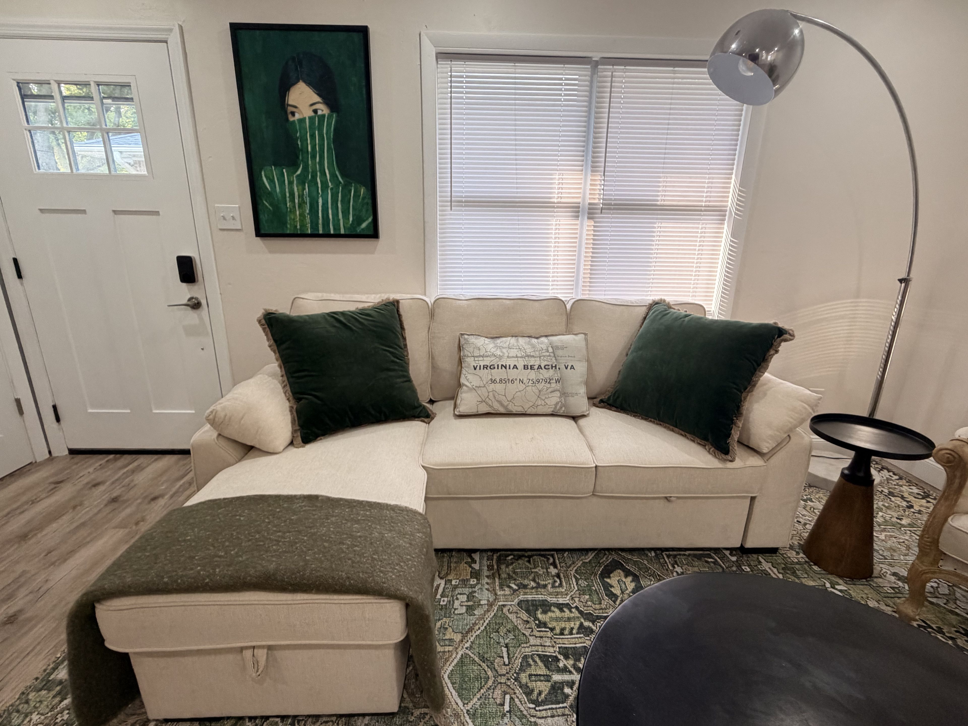 Un salon avec un canapé beige, deux coussins verts, un coussin décoré d’une carte, une lampe sur pied et un tapis à motifs.