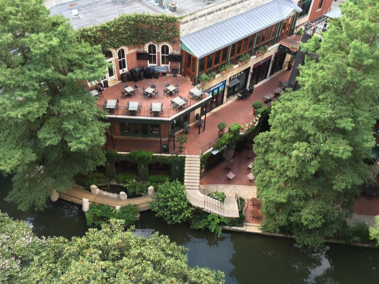 A riverside restaurant with outdoor seating, a bridge, and a building with a brick facade.