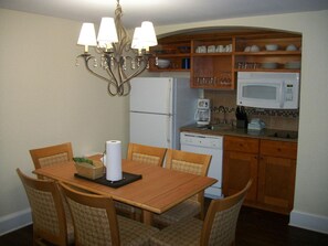 A dining area with a wooden table and chairs, a chandelier, a refrigerator, a microwave, and a kitchen counter with cabinets.