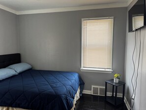 A bedroom with a bed, a window with blinds, a small table with a plant, and a wall-mounted TV.