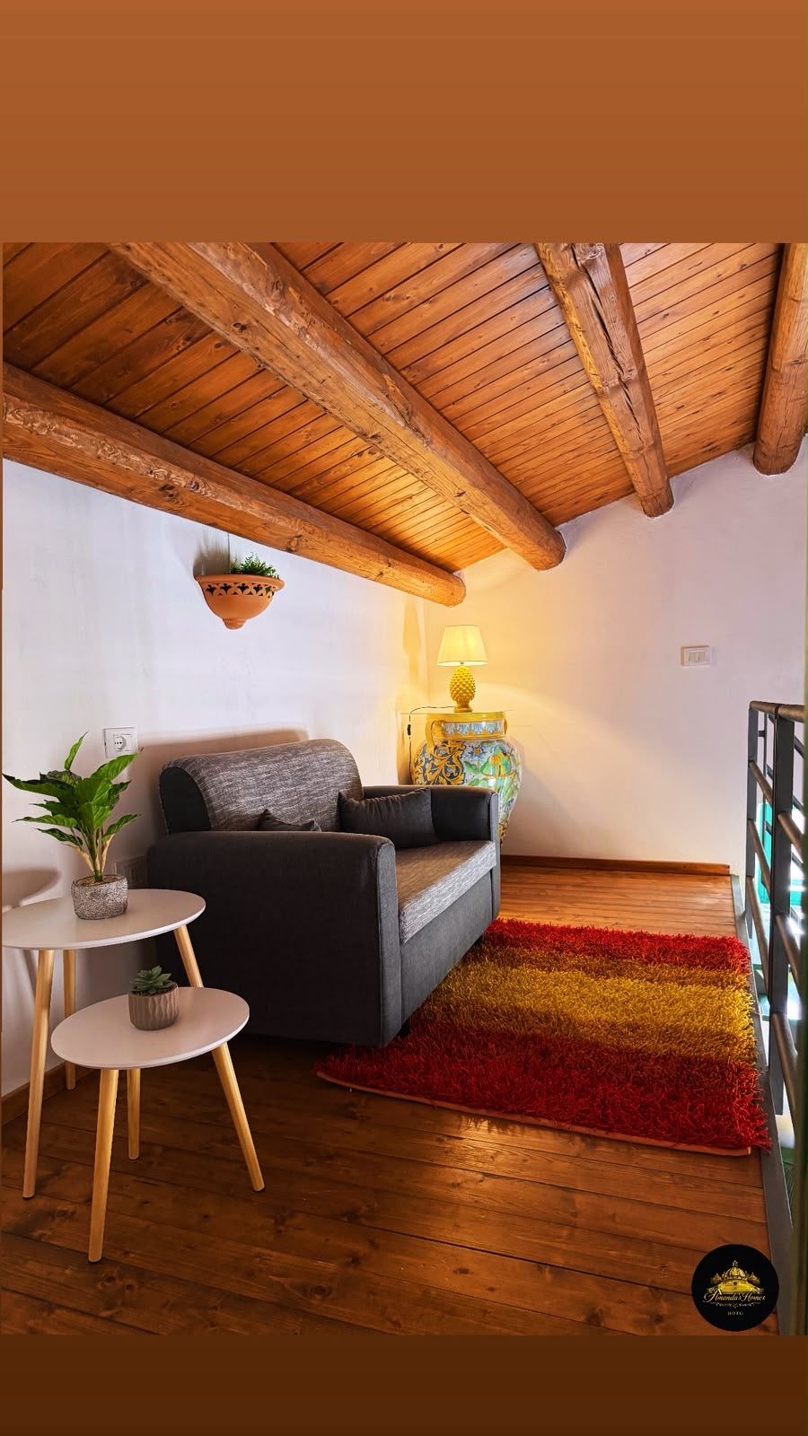Een gezellige woonkamer met een houten plafond, een bank, een klein tafeltje, een lamp en een kleurrijk vloerkleed.