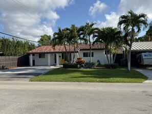 A single-story house with a red-tiled roof, a front yard with palm trees, and a clear sky. - The cozy little house. (Fort Lauderdale)
