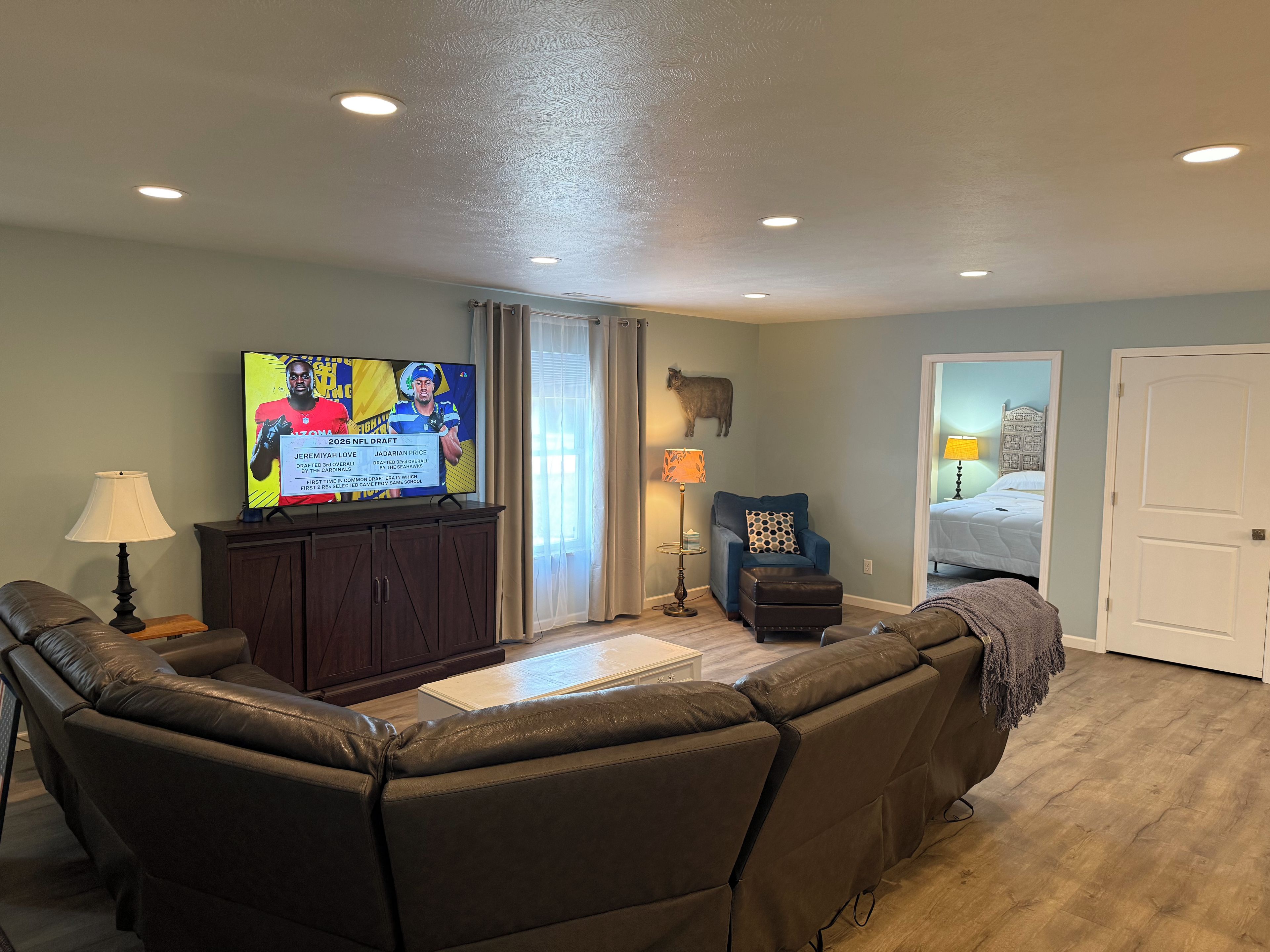 Una sala de estar con un sofá de cuero, un televisor mostrando contenido deportivo, una lámpara y un sillón azul.