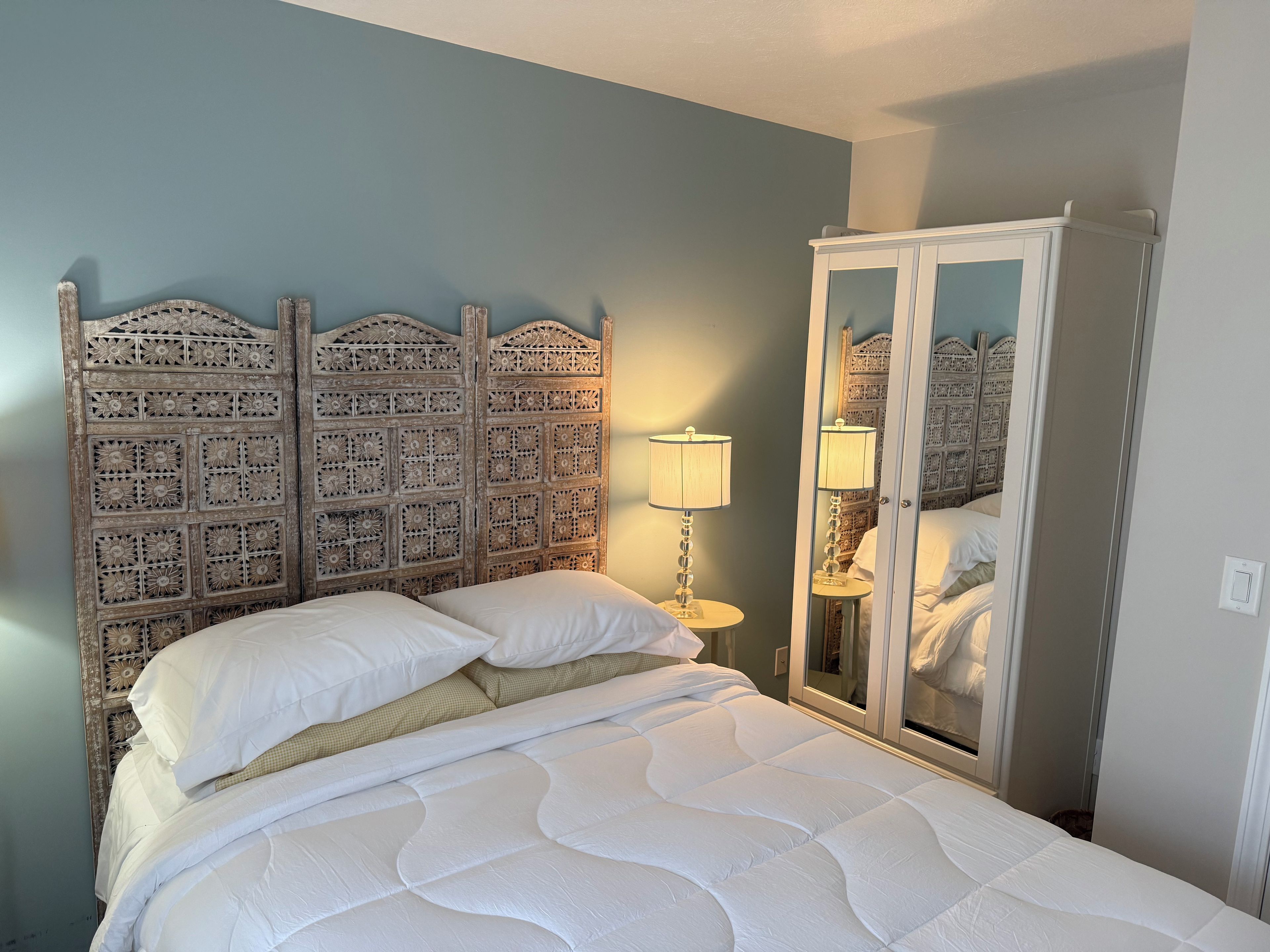 Ein Bett mit weißer Bettdecke, ein Holzkopfteil mit aufwendigen Mustern, eine Nachttischlampe und ein Kleiderschrank mit Spiegel.