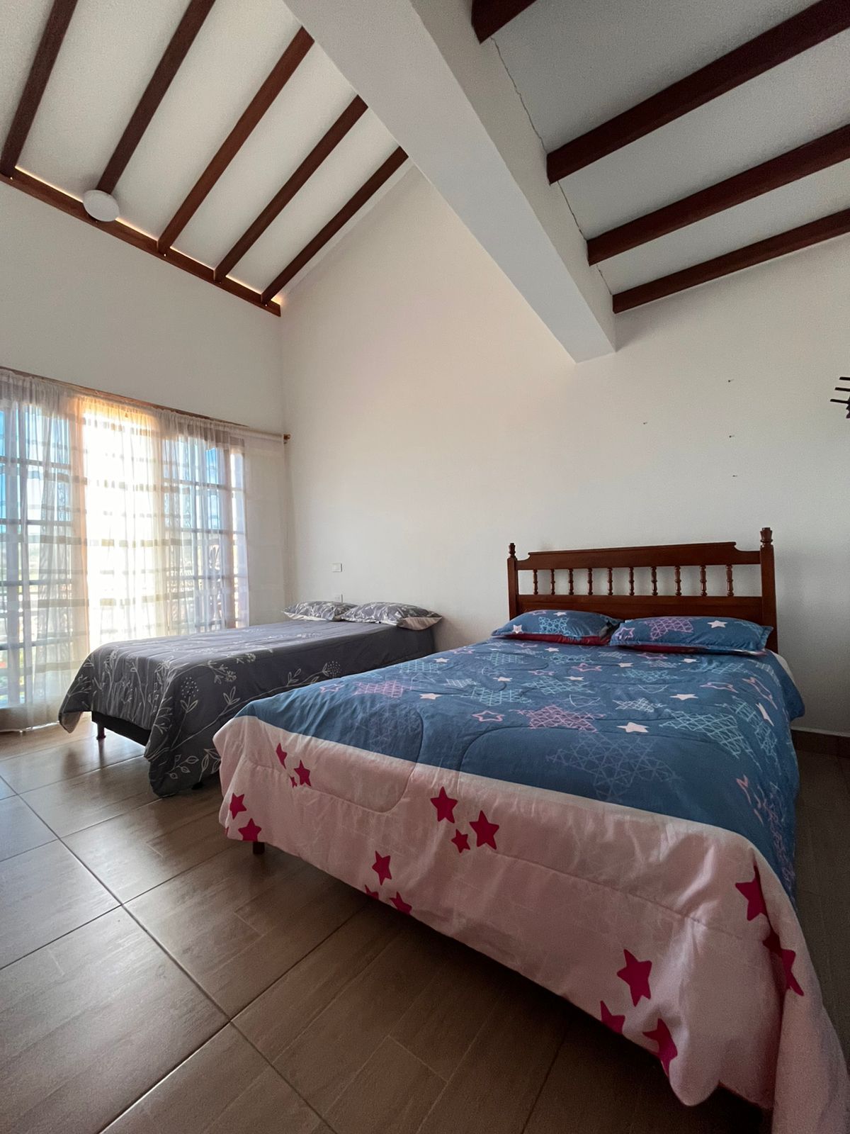 A bedroom with two beds, wooden beams on the ceiling, and a window with curtains.