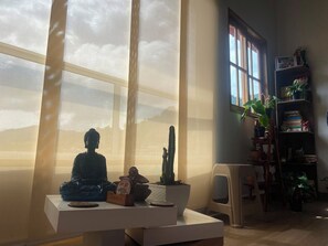 Ein Zimmer mit einem großen Fenster, einer Buddha-Statue, einem Kaktus und einem Bücherregal mit Pflanzen.