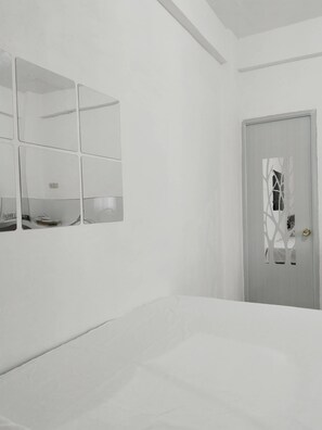 A minimalist bedroom with a bed, a wall-mounted mirror, and a door with a decorative pattern. - JNT Apartment is newly renovated studio with modern CR, w/ huge parking  space. (Pili)
