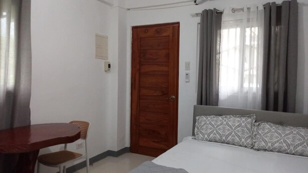 A bedroom with a wooden door, a bed with patterned pillows, a chair, and a table. - JNT Apartment is newly renovated studio with modern CR, w/ huge parking  space. (Pili)