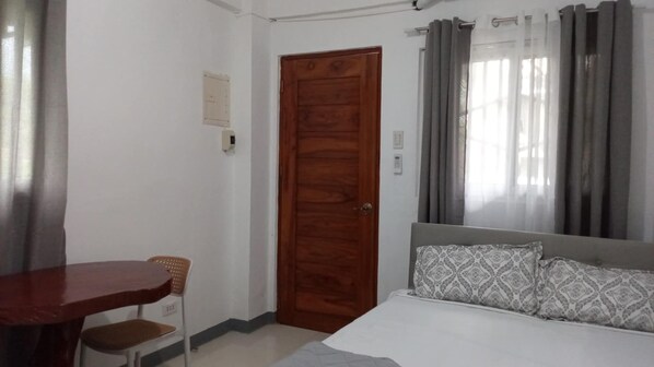 A bedroom with a wooden door, a bed with patterned pillows, a chair, and a table. - JNT Apartment is newly renovated studio with modern CR, w/ huge parking  space. (Pili)