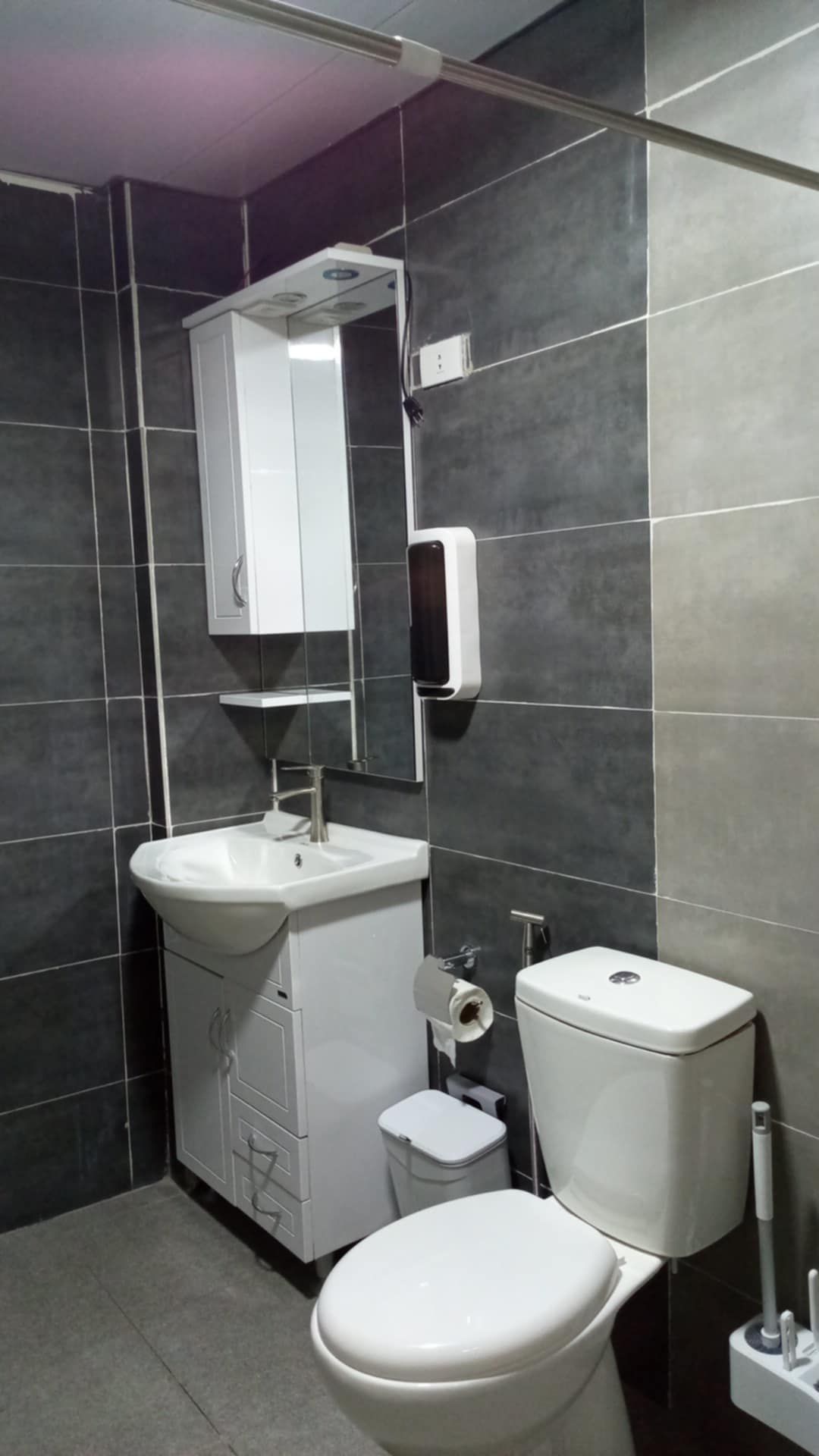 A modern bathroom with a white sink, mirror, toilet, and a small trash bin.