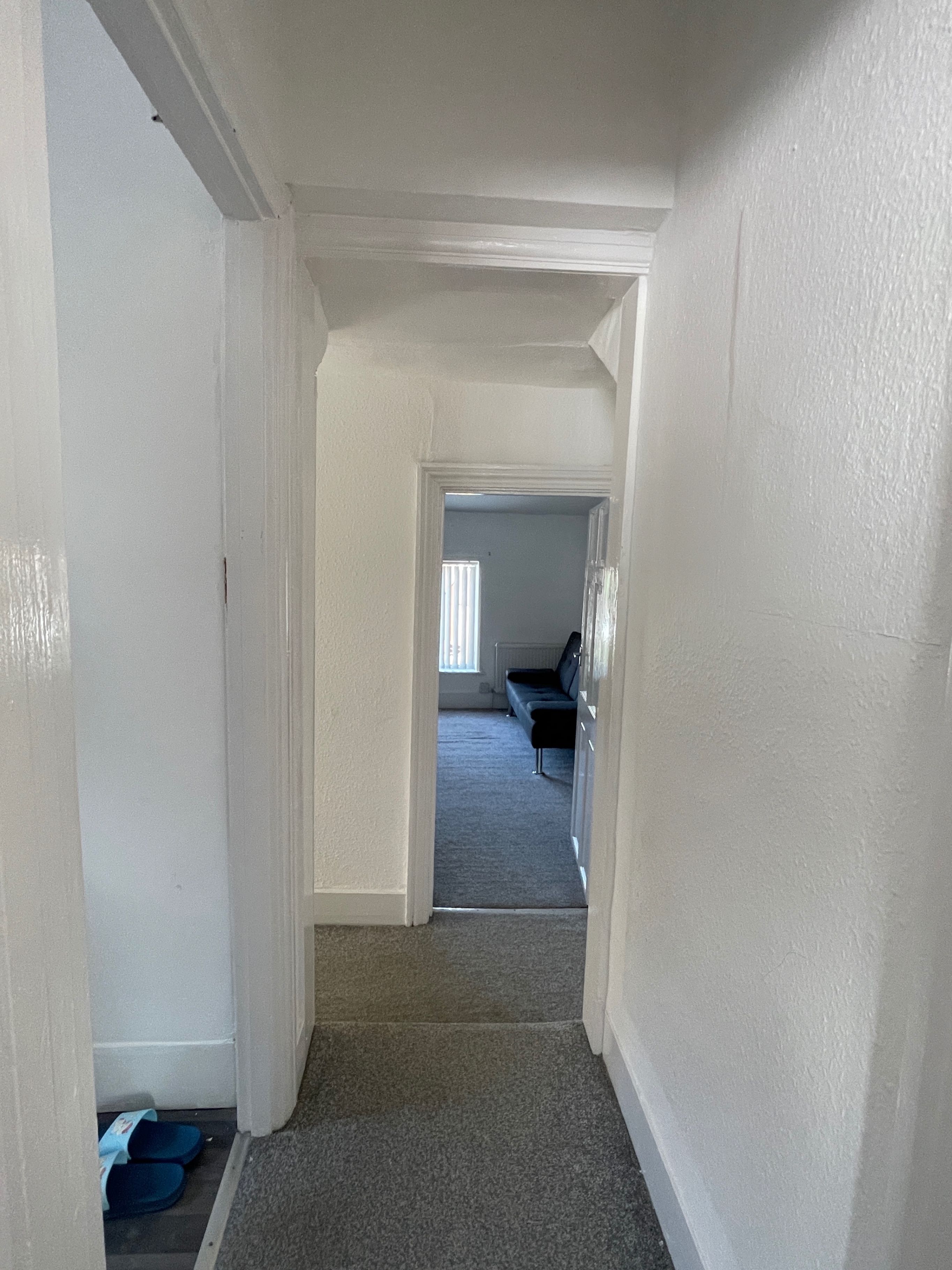 Een smalle gang met vloerbedekking die leidt naar een kamer met een raam en een bank.