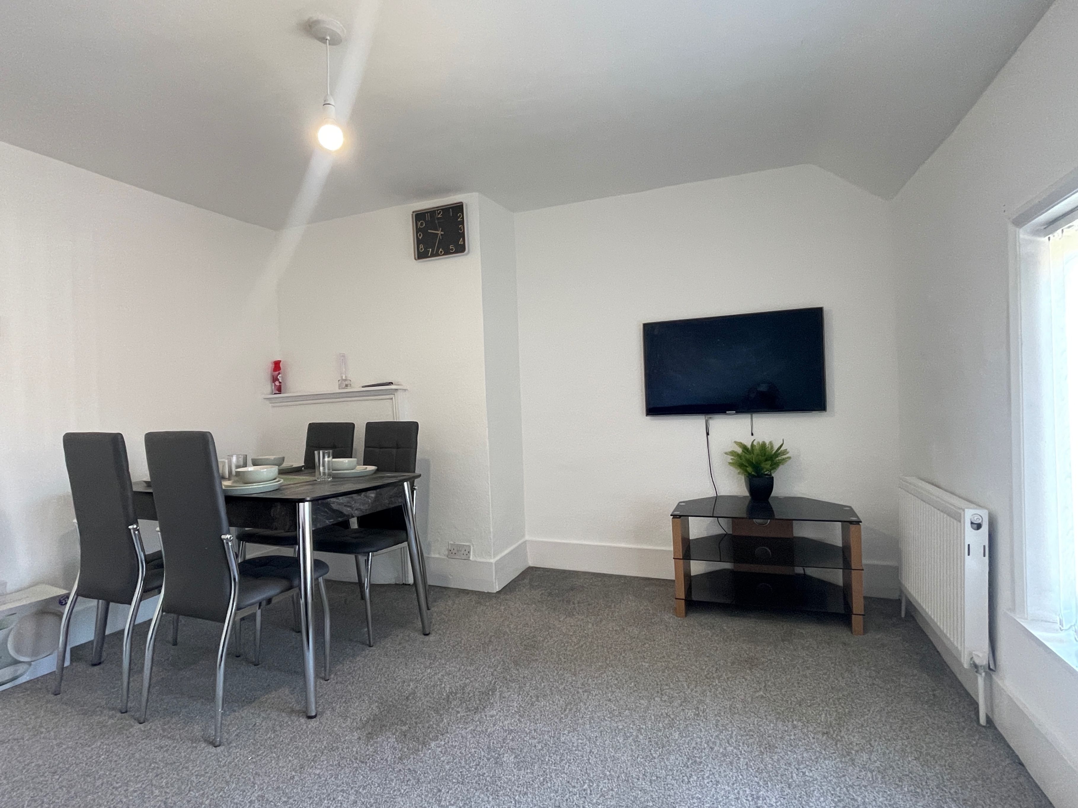 A room with a dining table, four chairs, a flat-screen TV mounted on the wall, a small plant on a stand, and a window with a radiator.